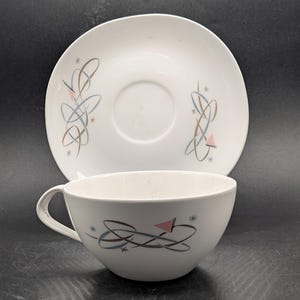 May include: A white ceramic teacup and matching saucer set featuring a mid-century modern pattern of intersecting brown and light blue loops, small pink triangles, and tiny starburst accents. The cup has a curved handle and a smooth finish.