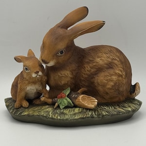 May include: A ceramic figurine of a mother rabbit and a baby rabbit. The rabbits are brown with detailed fur and are sitting on a green base. A small log with red berries and green leaves is between them.