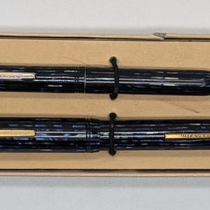 May include: A set of two blue and gold pens in a cardboard box. The pens have a striped pattern and the words "Sheaffer" and "USA" are visible on the pens.