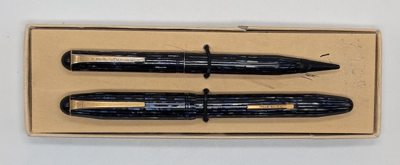 May include: A set of two blue and gold pens in a cardboard box. The pens have a striped pattern and the words "Sheaffer" and "USA" are visible on the pens.