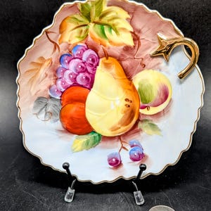 May include: A vintage porcelain fruit bowl with a gold rim and a handle in the shape of a vine. The bowl is decorated with a hand-painted design of pears, grapes, and peaches.