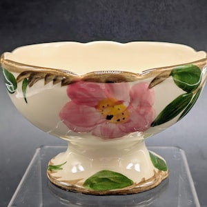 May include: Cream-colored pedestal bowl with a scalloped rim and a floral design. The bowl features a large pink flower with a yellow center, green leaves, and gold trim. A decorative piece for serving or display.