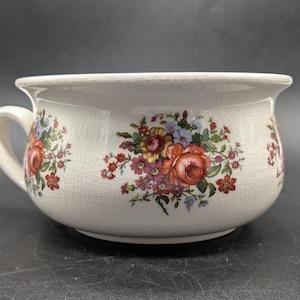 May include: A white ceramic chamber pot featuring a prominent crackle glaze finish and a decorative floral pattern. The design includes clusters of red, orange, yellow, and blue flowers with green leaves arranged around the exterior of the basin.