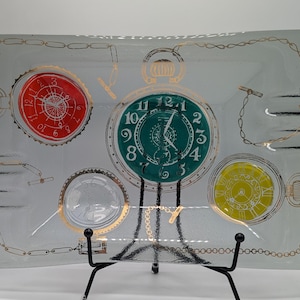 May include: Rectangular glass tray with a time-themed design. The tray features a clear background with gold-colored chain and pocket watch illustrations. The pocket watches are in red, green, and yellow. The tray is supported by a black metal stand.