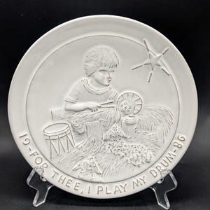 May include: White ceramic plate with a raised relief design. The design depicts a child playing a drum, with a baby and a star. The text around the edge reads "19-FOR THEE, I PLAY MY DRUM-86".