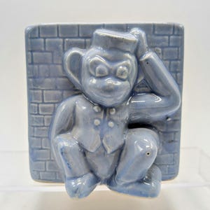 May include: A blue ceramic planter shaped like a monkey wearing a hat and a vest. The monkey is sitting against a brick wall.