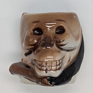 May include: A ceramic skull figurine with a brown and black painted design. The skull is wearing a black hat and has a brown pipe in its mouth.
