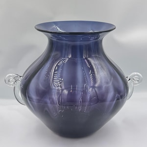 May include: A large, elegant, purple glass vase with a wide, flared rim and two clear glass handles. The vase has a rounded body and a dark purple base, with lighter shades of purple swirling upwards. The glass has a glossy finish.