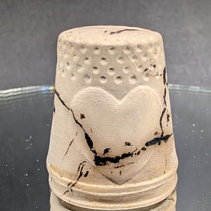 May include: A beige ceramic thimble with a heart design and dotted pattern. The thimble has black crackle detailing and is likely used for sewing. The thimble is approximately 1 inch tall.