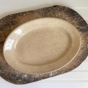 May include: An oval-shaped ceramic platter with a smooth, light beige glaze and a visible network of fine surface crazing. The platter features a slightly raised rim and a glossy finish, displaying subtle variations in color across its surface.