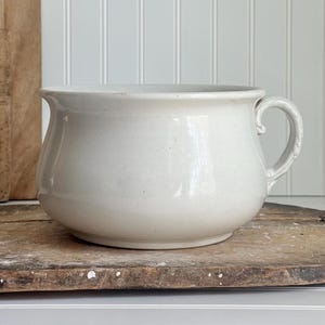 May include: Off-white ceramic bowl with a curved handle. The bowl has a rounded body and a slightly flared rim. It sits on a rustic wooden surface, with a wooden cutting board in the background. The bowl is likely for serving or display.