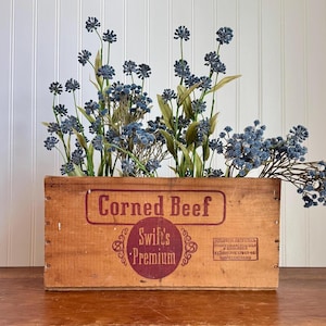 May include: A vintage wooden crate with the words "Corned Beef Swift's Premium" printed on the front. The crate is filled with blue flowers.