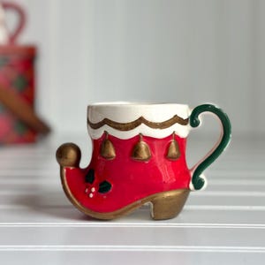 May include: A ceramic Christmas mug shaped like a red elf boot with a gold heel and toe, white trim, and a green handle. The boot features gold bells and holly accents.