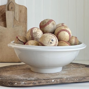 May include: A white ceramic bowl filled with vintage baseballs. The baseballs are off-white with red stitching and show signs of wear. The bowl sits on a wooden surface, with a wooden cutting board in the background.