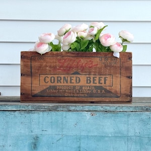 May include: A rustic wooden box, originally for corned beef, now filled with pink and white flowers. The box has faded red lettering and the words "CORNED BEEF" prominently displayed. The box is sitting on a weathered blue surface.