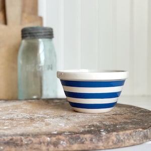 May include: A small, off-white ceramic bowl with horizontal blue stripes. The bowl has a slightly flared rim and sits on a rustic, wooden surface. A vintage-style glass jar is visible in the background.