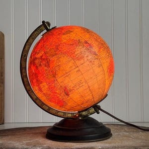 May include: A spherical illuminated globe featuring a warm orange and red glow, displaying detailed world map cartography. The globe is mounted within a dark metallic meridian ring and rests on a circular black base, with a power cord extending.