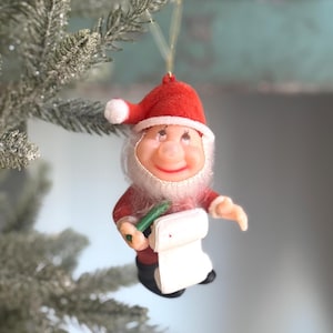 May include: A vintage Christmas ornament shaped like Santa Claus, wearing a red felt hat and suit with a white beard. The ornament is holding a green pen and a white list. The ornament is hanging from a silver Christmas tree.