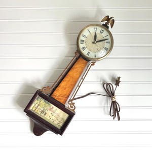 May include: Vintage wall clock with a gold-tone face, Roman numerals, and a decorative eagle on top. The clock has a wooden frame with a picture of a building and a brown power cord. The clock is approximately 12 inches tall.