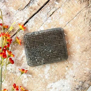 May include: A rectangular, gray flower frog with numerous metal pins. The frog is made of a heavy material and is designed to hold flower stems in place. The background is a weathered, light-colored wooden surface.