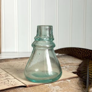 May include: A clear, antique-style glass bottle with a rounded base and a flared neck. The bottle has a slight teal tint and sits on a vintage ledger. The bottle's design is reminiscent of apothecary or vintage perfume bottles.