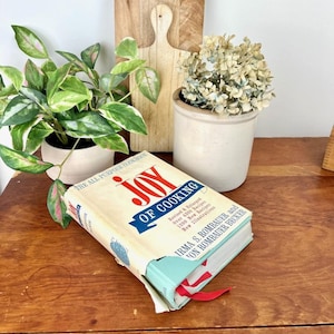 May include: A vintage cookbook titled "Joy of Cooking" with a light blue spine and a red ribbon bookmark. The book is placed on a wooden surface, next to a small potted plant with green and pink leaves and a small pot of dried flowers.