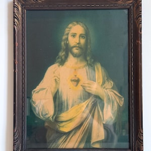 May include: A framed religious print featuring a portrait of Jesus Christ. The image shows Jesus with long hair, a beard, and a glowing heart. The frame is dark brown with ornate detailing, adding a vintage aesthetic to the artwork.