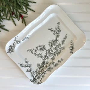 May include: A white rectangular serving tray with a curved edge, decorated with a black and gray floral branch design. The tray is placed on a white surface, with a sprig of greenery in the upper left corner.