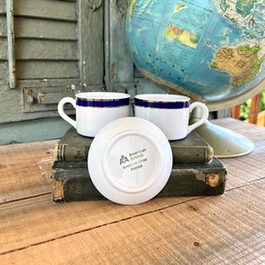 May include: Two white ceramic coffee cups with blue and gold trim, and a saucer with the words "American Airlines Syracuse China 730159" printed on the bottom. The cups and saucer are sitting on top of a stack of old books.
