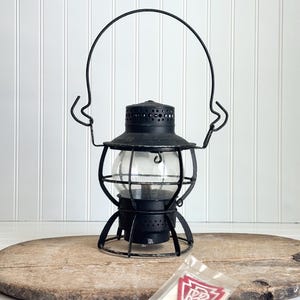 May include: A black vintage lantern with a clear glass globe and a wire handle. The lantern sits on a wooden surface. A small, clear plastic bag with a red and white patch is in the foreground. The patch has the letters "PRR" inside a white shape.