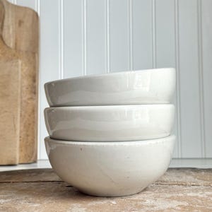 May include: A stack of three off-white ceramic bowls with a smooth, glossy finish. Each bowl features a rounded shape and a consistent, neutral color palette. The bowls are stacked vertically, showcasing their uniform design and simple, clean edges.