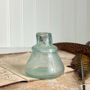 May include: A small, antique, light blue glass inkwell with a rounded base and a flared neck. The inkwell sits on a vintage paper with handwritten text. A brown and yellow feather is next to the inkwell.