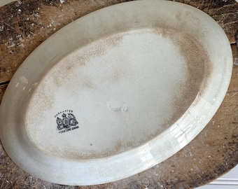 May include: An oval-shaped off-white ceramic platter with a visible crackle glaze finish. The underside features a black printed stamp with a crown and unicorn emblem, including the text WARRANTED IRONSTONE CHINA.