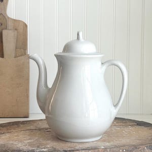 May include: A white ceramic teapot with a lid and handle. The teapot has a curved spout and a rounded body. The background includes a wooden cutting board and a white wall with vertical paneling.