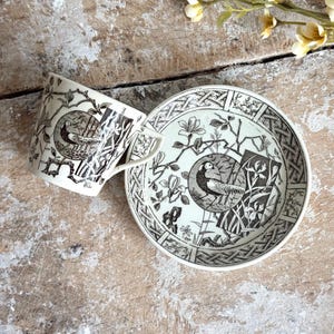 May include: A cream-colored ceramic teacup and saucer set featuring a black transferware pattern of pheasants within circular frames, surrounded by botanical illustrations of flowers, branches, and geometric lattice borders on both pieces.