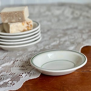 May include: A white ceramic dish with a green rim. The dish is empty and sitting on a white lace tablecloth.