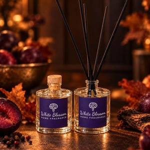 May include: Two clear glass bottles of White Blossom Home Fragrance with dark blue labels featuring a tree icon. One bottle has a cork stopper, while the other contains five black reed diffuser sticks inserted into the neck of the bottle.