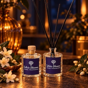 May include: Two clear glass bottles with dark blue labels featuring a white tree icon and the text White Blossom HOME FRAGRANCE. One bottle has a light wood stopper, and the other contains five thin black reed diffuser sticks.