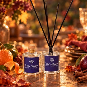 May include: Two clear glass bottles with cork stoppers and dark blue labels featuring a white tree icon and the text White Blossom HOME FRAGRANCE. One bottle contains black reed diffuser sticks, while the other is sealed with a cork.