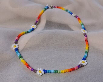 May include: A rainbow beaded necklace with white daisy flower accents. The necklace is made with small colorful beads and features a simple design.