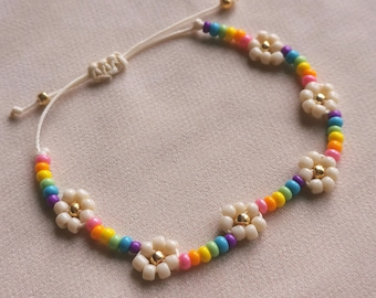 May include: A colourful beaded bracelet with white flower charms and gold accents. The bracelet is made with rainbow coloured beads and has a white string closure.