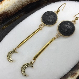 May include: A pair of gold earrings featuring black lava rock and crescent moon charms. The earrings are dangle style and have a simple, minimalist design.