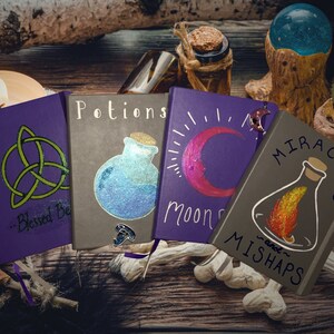 May include: Four purple and gray journals with different designs and titles. The journals are on a wooden surface with a candle, a glass orb, and bones. The titles of the journals are "Blessed Be", "Potions", "Moon Spells", and "Miracles and Mishaps".