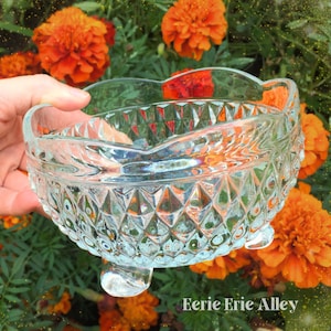 May include: A clear, heart-shaped glass bowl with a diamond pattern and three small feet. The bowl is held in a hand against a backdrop of orange marigold flowers and green foliage. The text "Eerie Erie Alley" is visible.