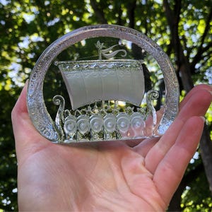Pode incluir: Um ornamento de vidro transparente em forma de semicírculo, apresentando um design gravado de um navio viking com uma vela grande e uma fileira de escudos na lateral. A borda externa do vidro tem um acabamento fosco e texturizado.