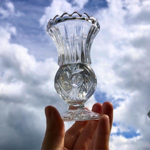 May include: A clear, ornate glass candlestick holder with a scalloped rim and a round, patterned base. The candlestick is held up against a cloudy blue sky, highlighting its intricate design and transparency. The base has a diameter of approximately 5 cm.