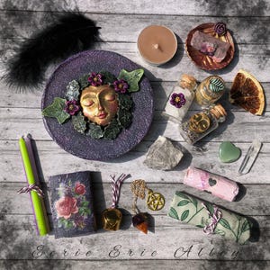 May include: An assortment of spiritual and metaphysical items, including a purple plate with a face design, candles, crystals, and small glass bottles. The image also features a small book with a floral design and a gold-colored pendant.