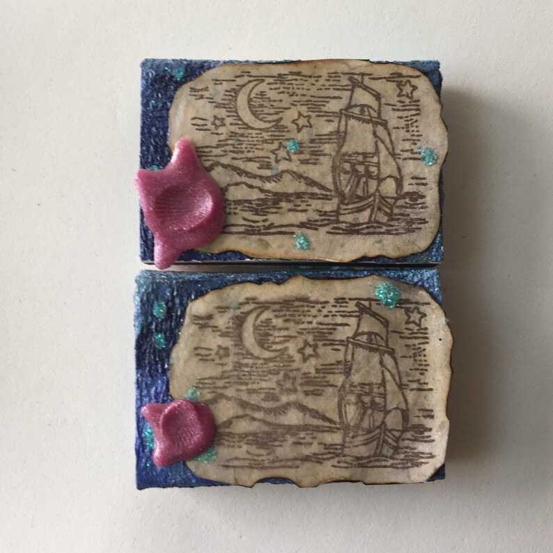 May include: Two small square canvases with a blue background and a brown paper image of a ship sailing under a crescent moon and stars. Each canvas has a pink 3D star on the left side.