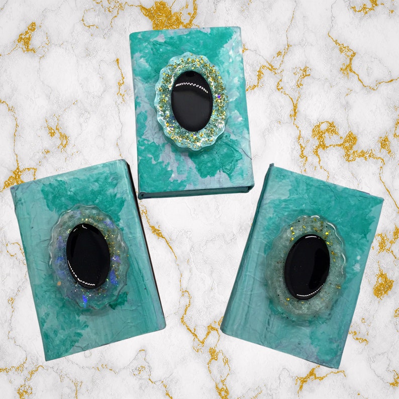 May include: Three teal-colored, decorative boxes with oval, black centerpieces. The centerpieces are framed by a light blue, scalloped border with gold glitter accents. The boxes are arranged on a marble-patterned surface.