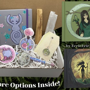 Puede incluir: Una caja blanca llena de varios artículos, incluyendo un pequeño cuaderno gris con un diseño floral y las palabras "Blessed Be" en la portada, una pequeña botella de líquido con un diseño de flor rosa, un palo de madera, un cristal transparente, un collar con un colgante de árbol, una pequeña vela, un lápiz y dos pequeños cuadernos con portadas pintadas. Los cuadernos presentan a una mujer con cuernos y un bastón de pie frente a una luna creciente, y a una mujer con un bastón de pie frente a un sol.